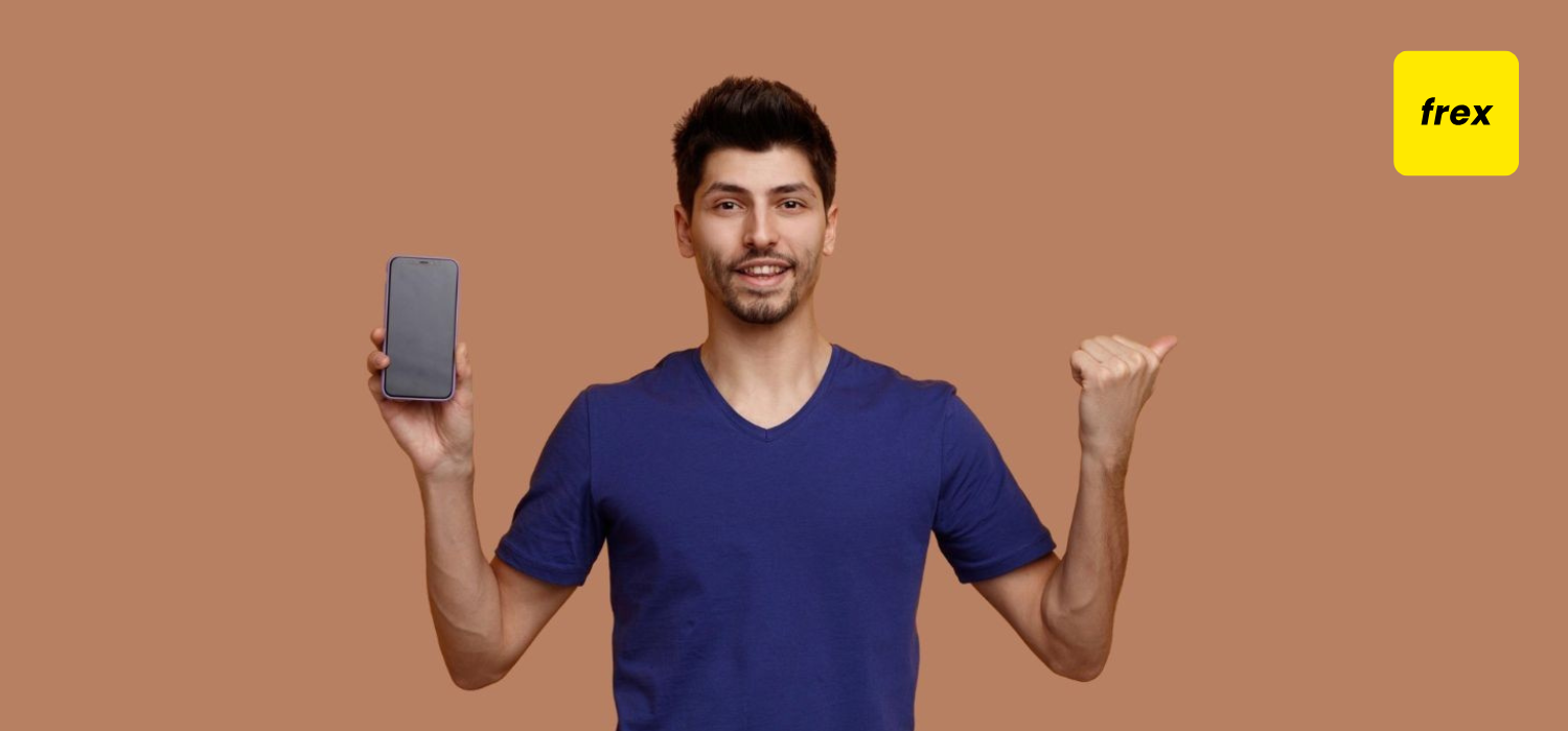 Smiling young handsome man looking at camera showing mobile phone to camera pointing to side