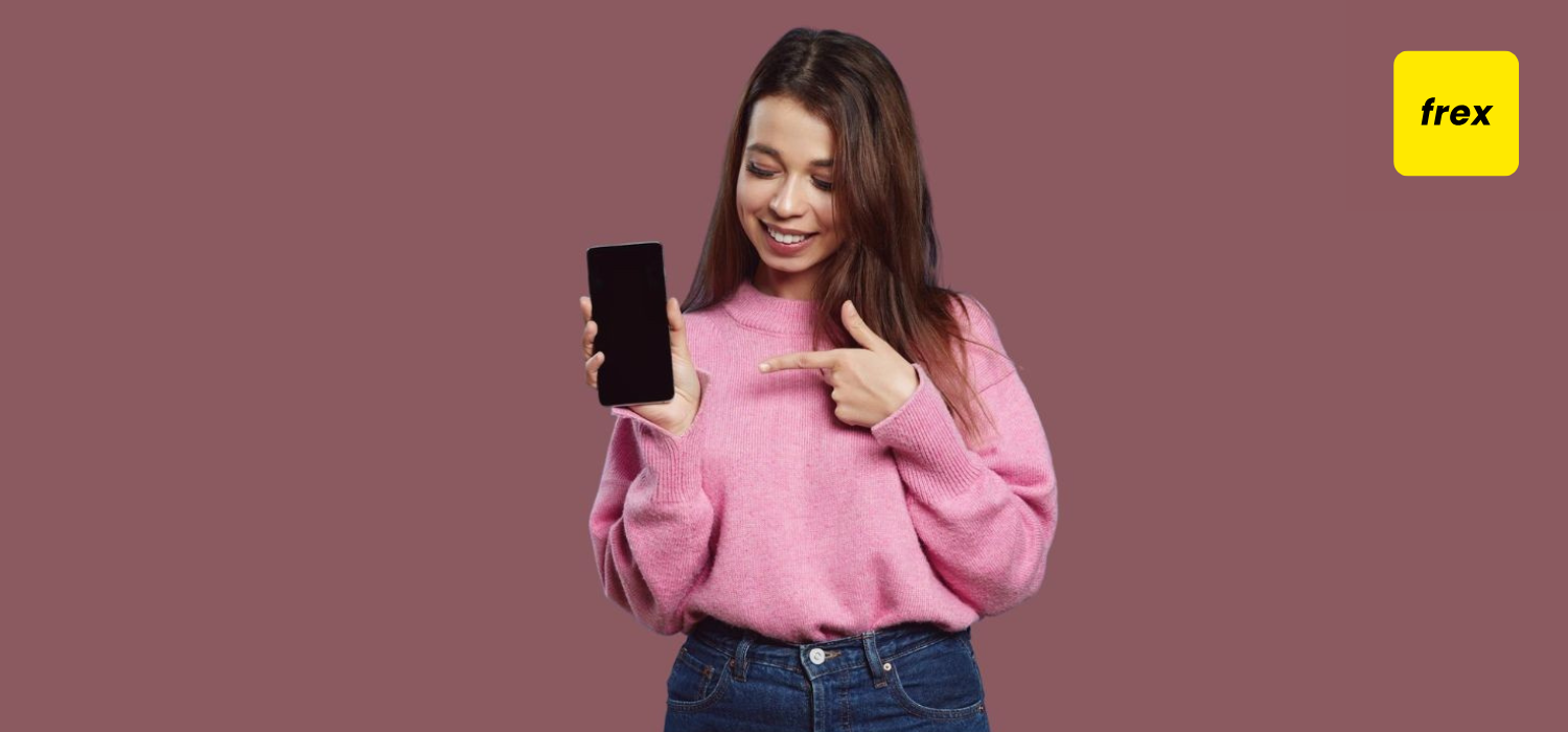 Happy young female in pink sweater smiling and looking at cellphone while pointing finger to mobile